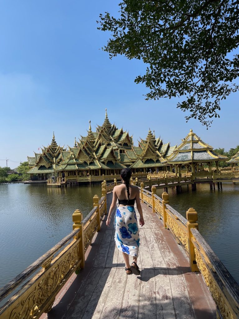 A woman walking on a beautifully structured bridge that leads to a gold and green temple complex over a lake.
