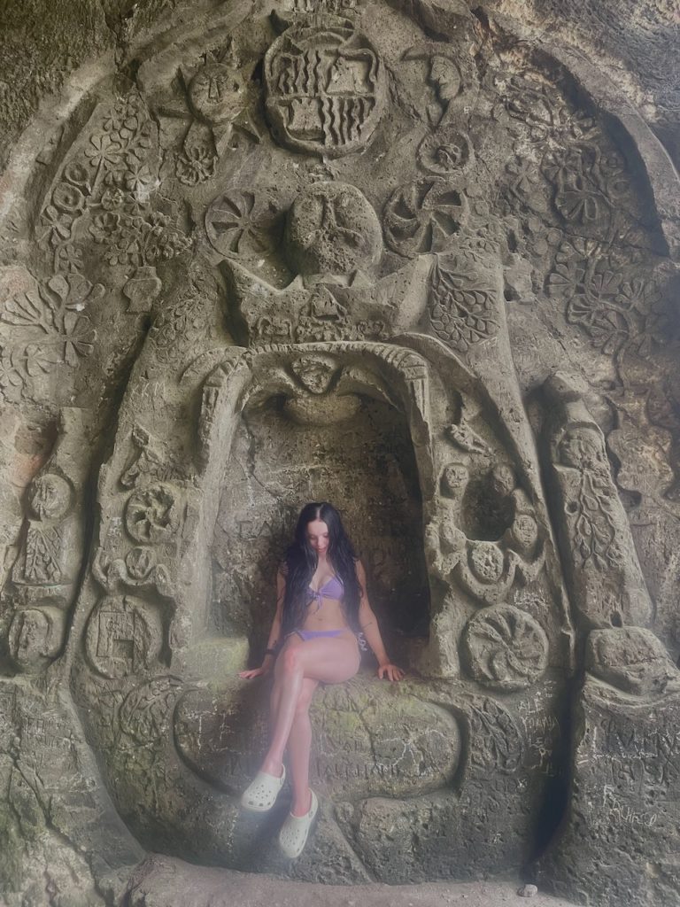 A woman seated within a cave with unique carvings surrounding her.