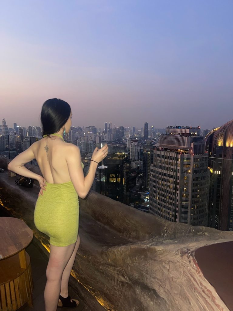 A woman facing the skyline of Bangkok with dozens of skyscrapers, with a drink in her hand.