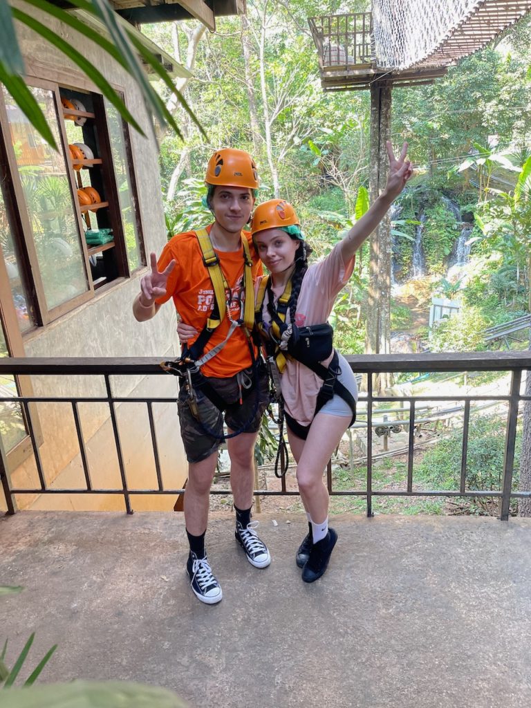 A couple in orange helmets and harnesses, ready to undertake ziplining in a jungle.