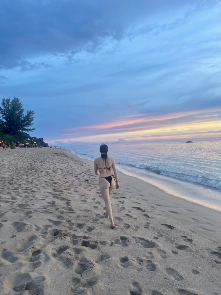 A woman in a black bikini strolling on the beach, enjoying the pastel sunset barefoot in the soft sand.