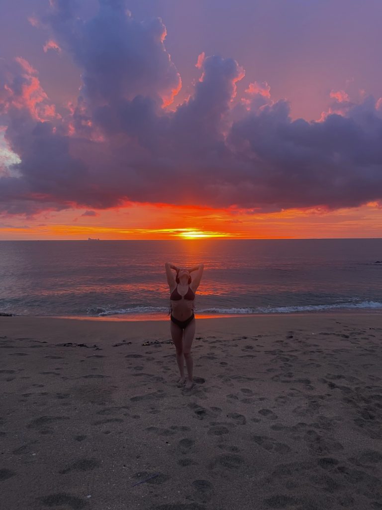 A woman joyfully raises her arms while standing on the beach, embracing the beauty of the surroundings at sunset.