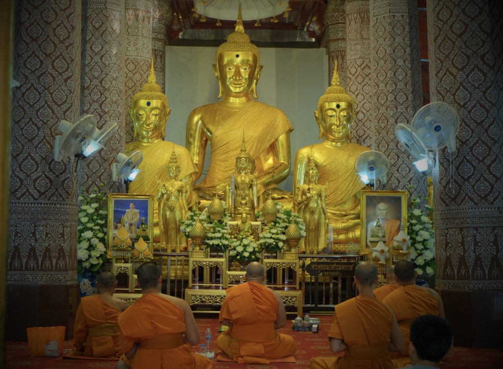 Monks in orange robes gathered in front of 3 large Buddha statues in a temple.