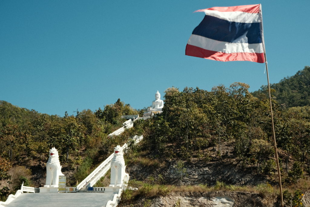 A big white Buddha situated on the top of a hill, with a Thai flag waving in the wind.