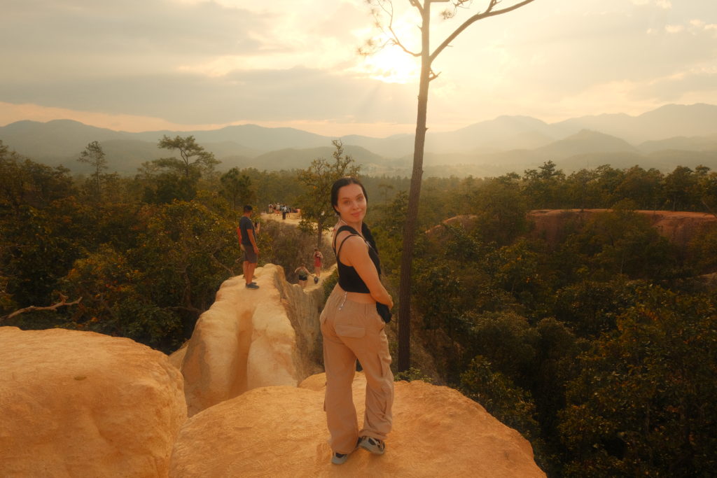 A woman standing on a canyon with majestic mountains towering in the background, at sunset.