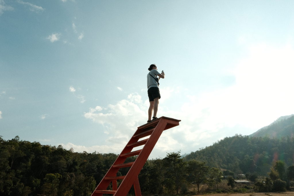A man standing on a tall red ladder with a dead drop, overlooking the rice fields.