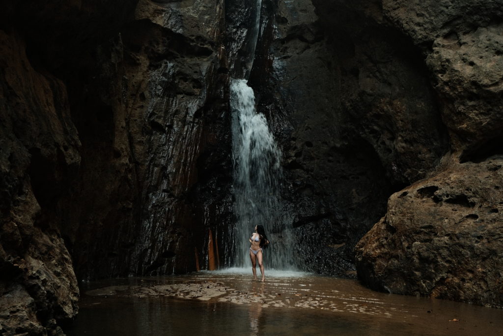 A woman stood beneath a free flowing waterfall, surrounded by huge rocky terrain.
