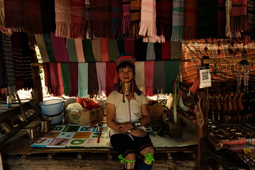 A 'Long Neck Karen' tribe woman smiling in front of her stand of handmade scarves.