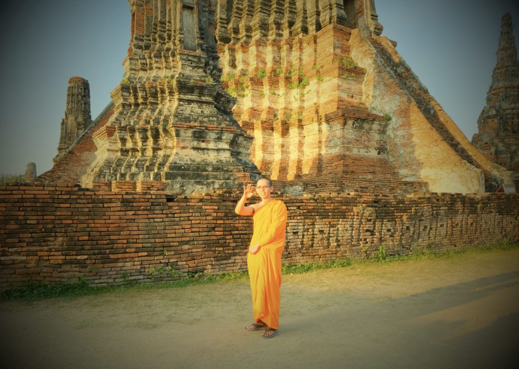 A young monk dressed in long orange robes, waving at the camera with a beautiful ancient temple in the background.