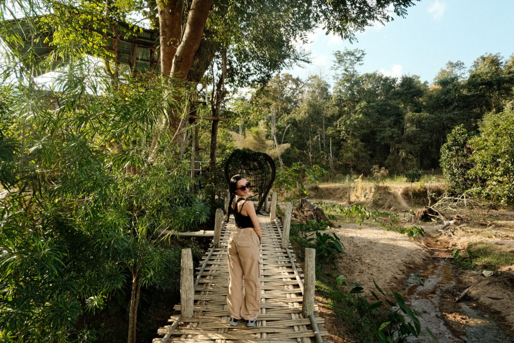 A woman standing on a locally crafted bamboo bridge, surrounded by green fields.