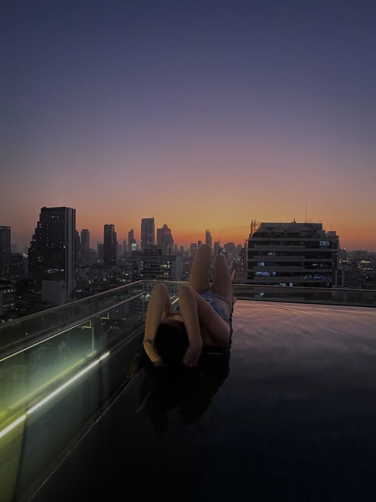 A woman posing on the edge of an infinity pool overlooking incredible city views and a hazy sunset.