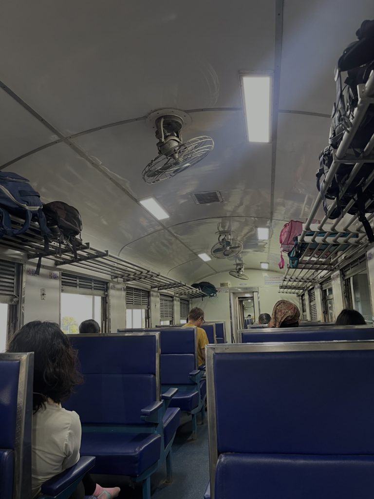 A glimpse inside a 3rd class train in Thailand with blue benches and luggage racks.