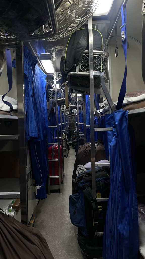Bunk beds inside a sleeper train with blue curtains and luggage racks.