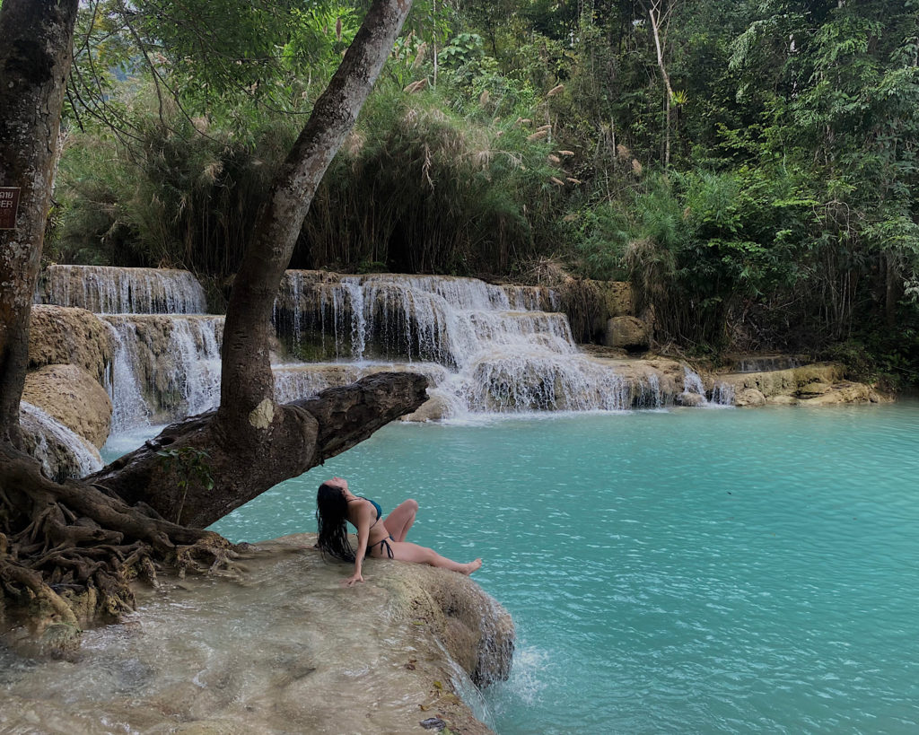 A woman peacefully sits on a rock near a cascading waterfall, enjoying the serenity of the natural area.