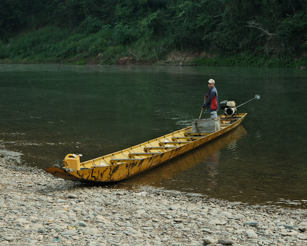 A long but narrow yellow boat on dark green waters, surrounded by greenery.