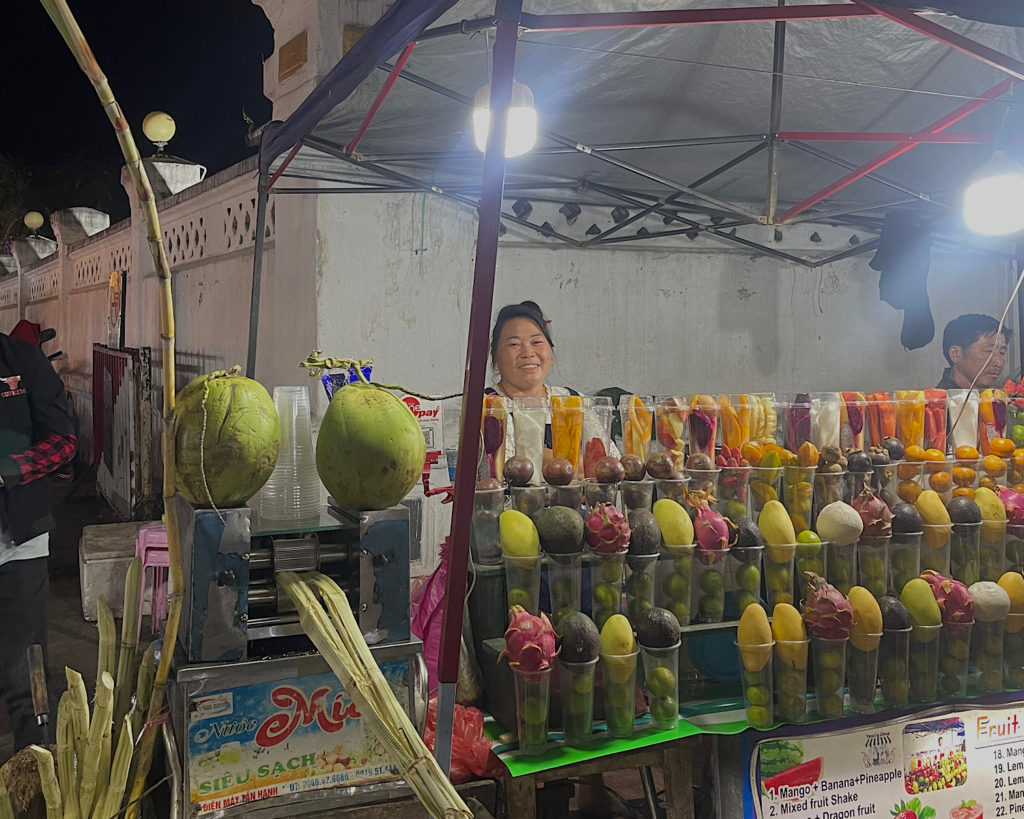 A friendly woman smiles behind a smoothie stall.