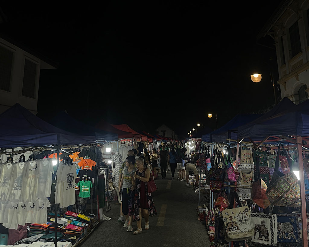 A bustling street market illuminated by lighting, with vendors selling various goods under the night sky.
