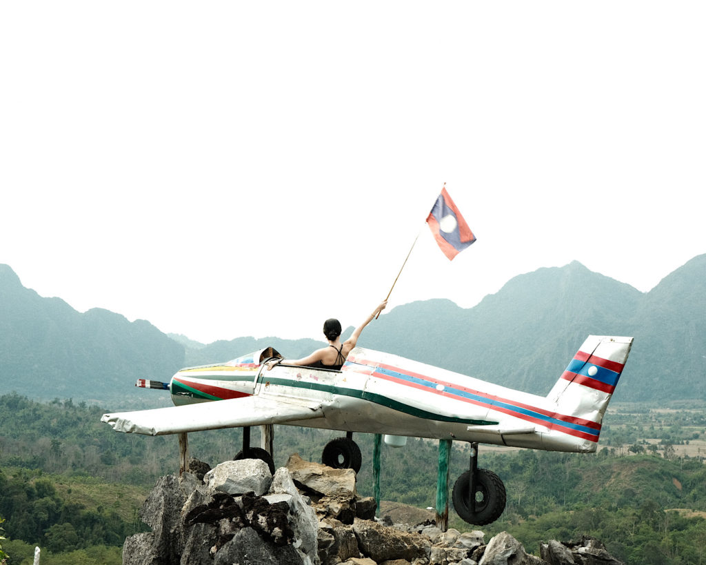 A woman seated in a makeshift plane, on top of a cliff, holding the Laos flag.