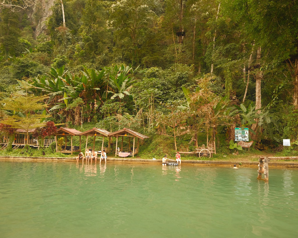 A blue lagoon surrounded by green tropical plants and wooden huts.