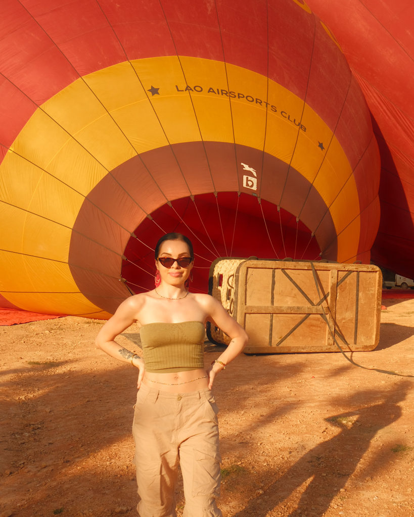 A woman posing in front of a partially inflated hot air balloon and basket resting on the floor.