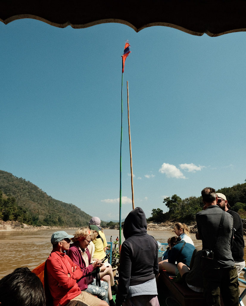 A group of passengers on the front of a wooden slow boat in Laos on a bright sunny day.