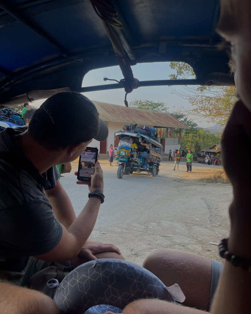 An overloaded tuk tuk carrying many passengers and a lot of backpacks on its roof.