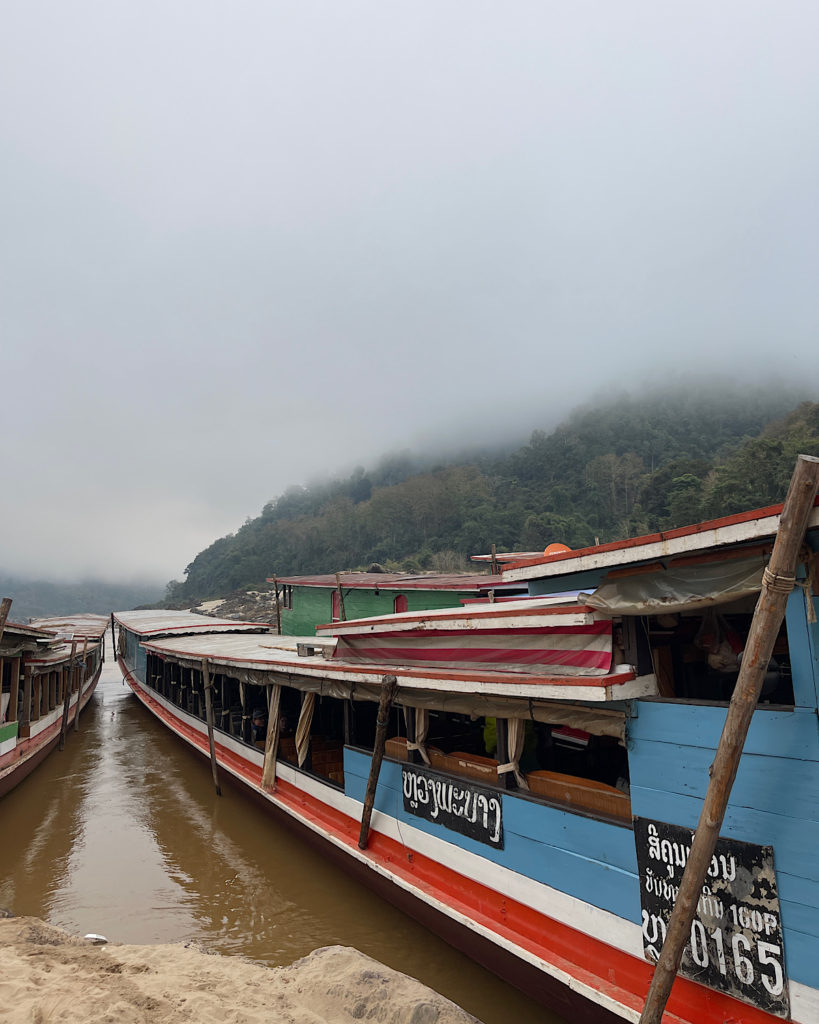Blue and red long wooden boats docked on the side of the Mekong River on a magical misty morning in Pakbeng.