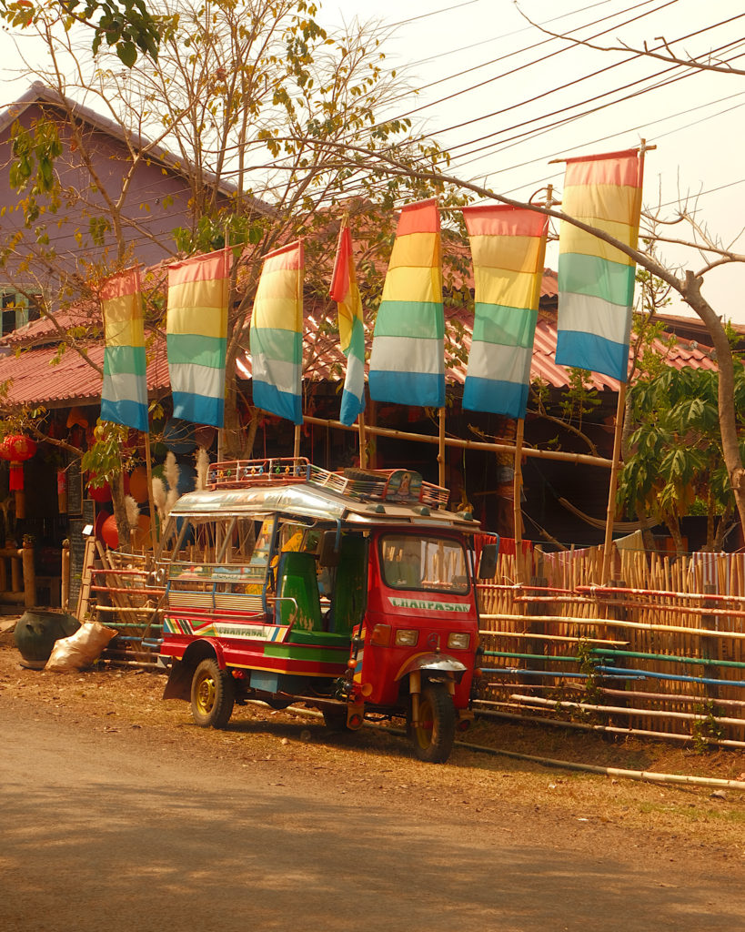 A multi coloured tuk tuk parked on the side of a dirt road, with colourful flags parked behind it.