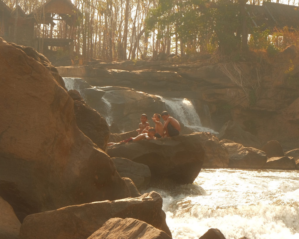 Three local children sat on a rock in front of a waterfall in nature.