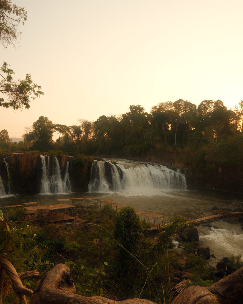 A majestic waterfall, on the Pakse Loop, cascading down a rocky cliff, surrounded by lush greenery at sunset.