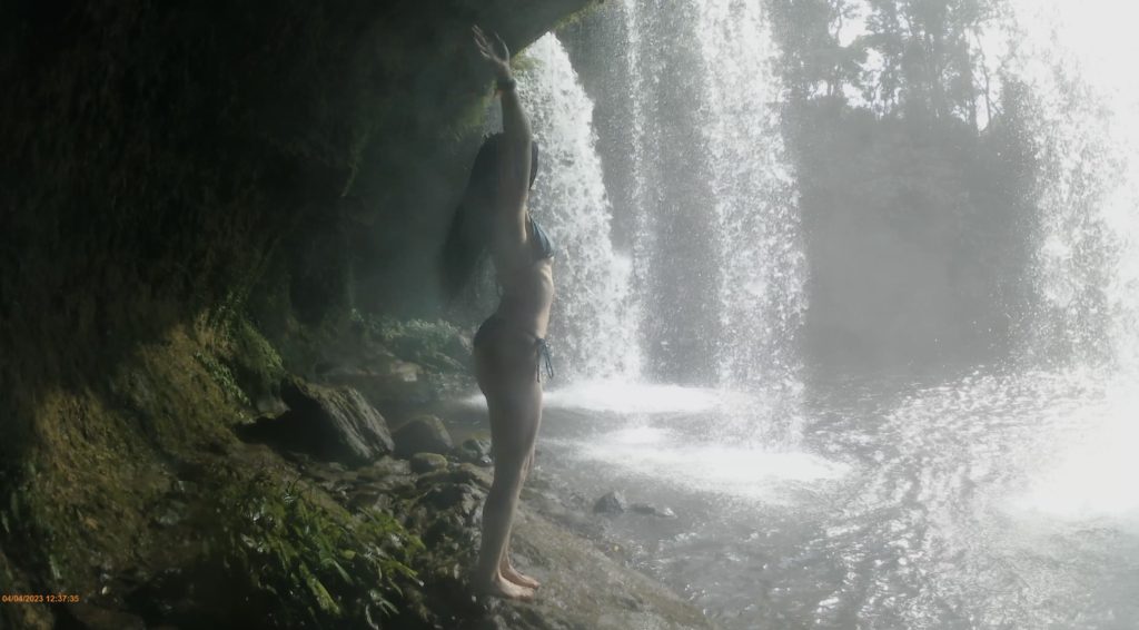 A woman in a bikini, stood behind a flowing waterfall, surrounded by lush greenery.