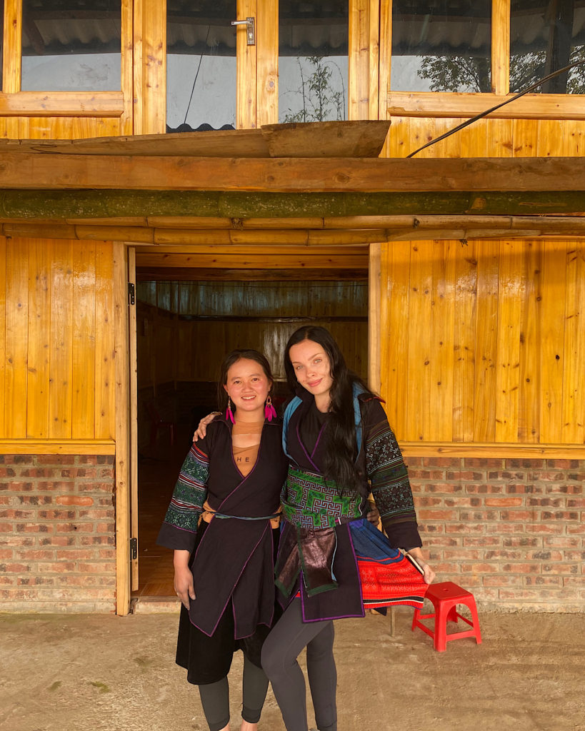Two women stood next to each other, dressed in Vietnamese ethnic outfits, outside a wooden house.