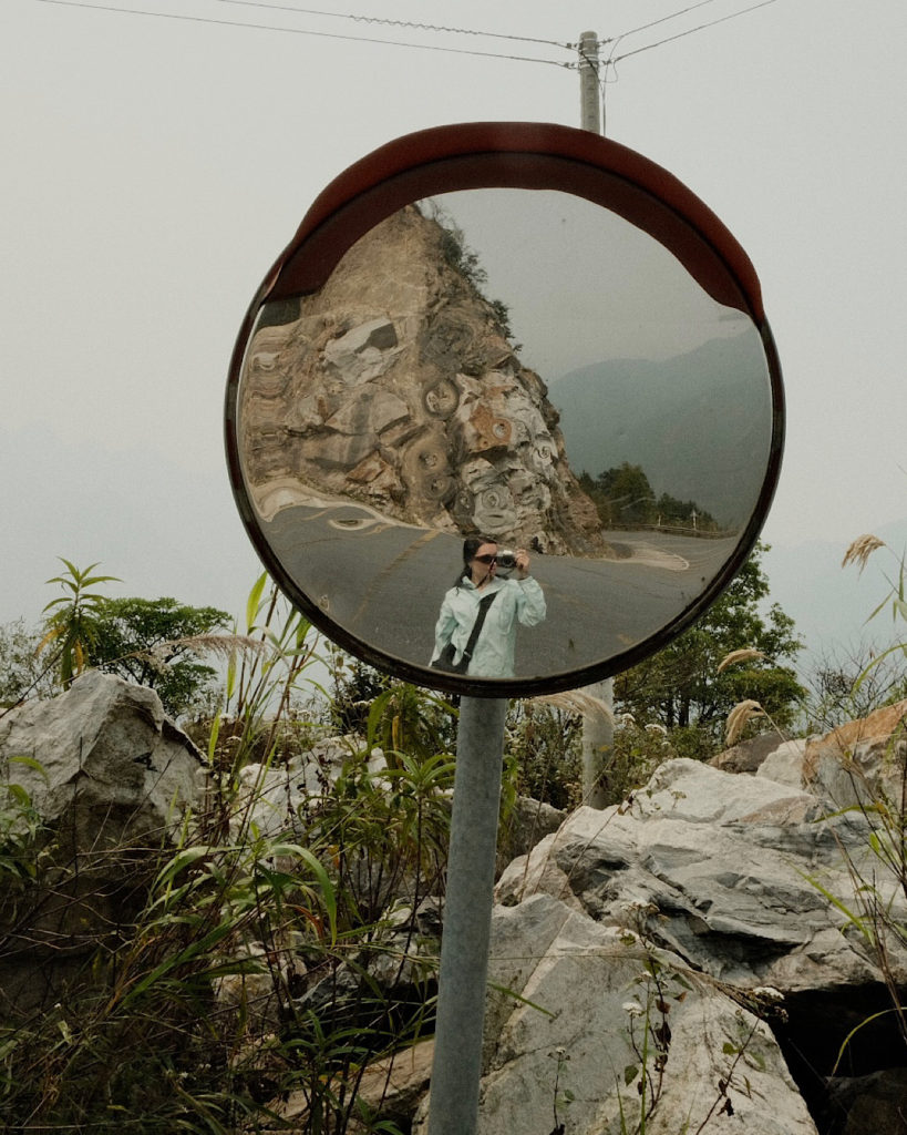 A person standing in front of a mirror on the roadside, surrounded by mountains and rice fields.
