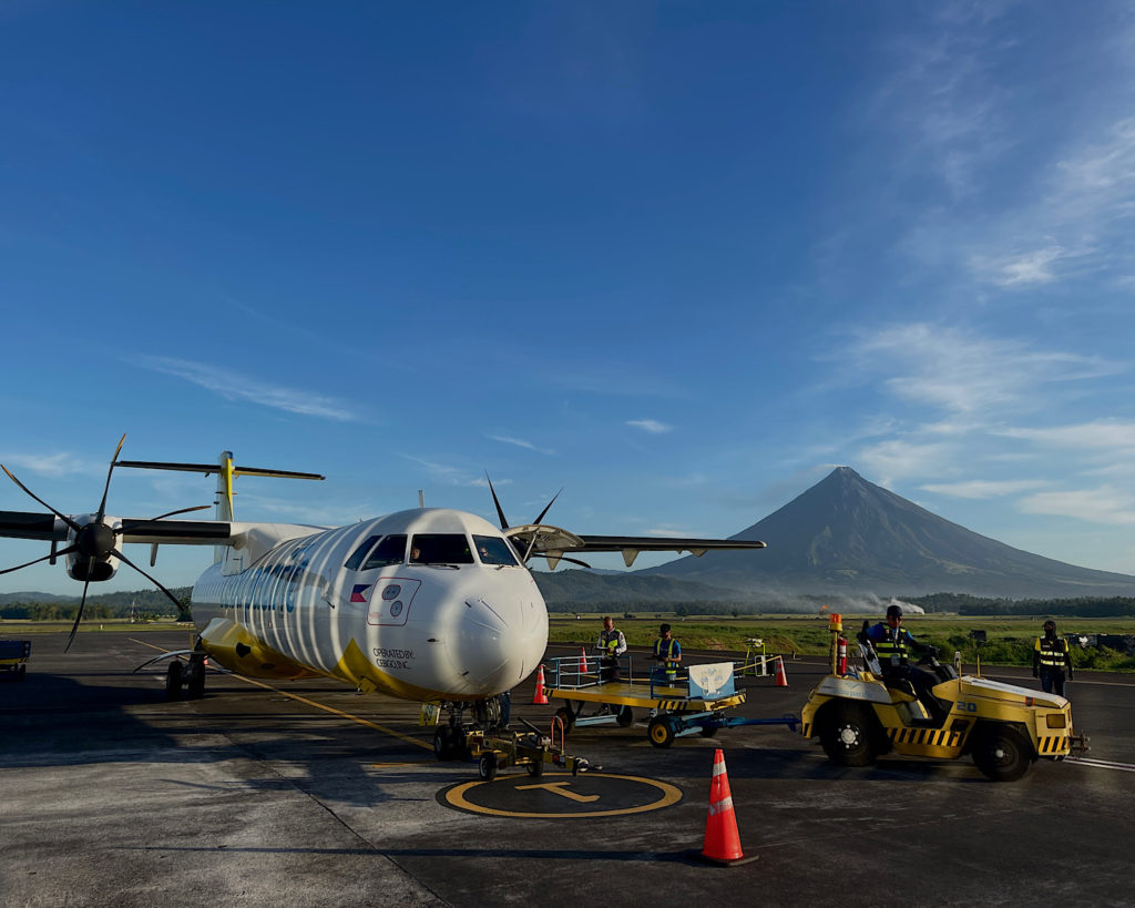 A white and yellow plane stationed on the runway, with a huge volcano in the background.