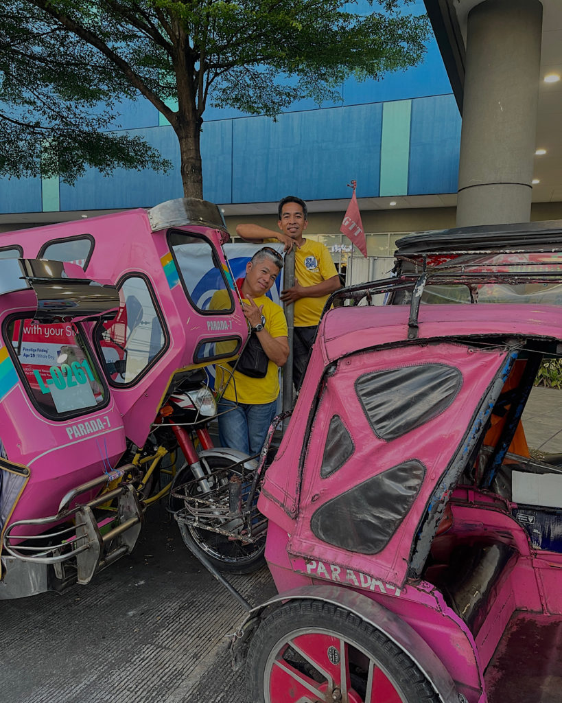 Two friendly Filipinos waving at the camera in front of their pink tricycles.