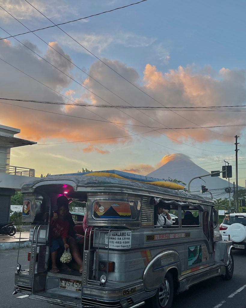 A silver jeepney driving through the city at sunset, with a cone volcano in the background.