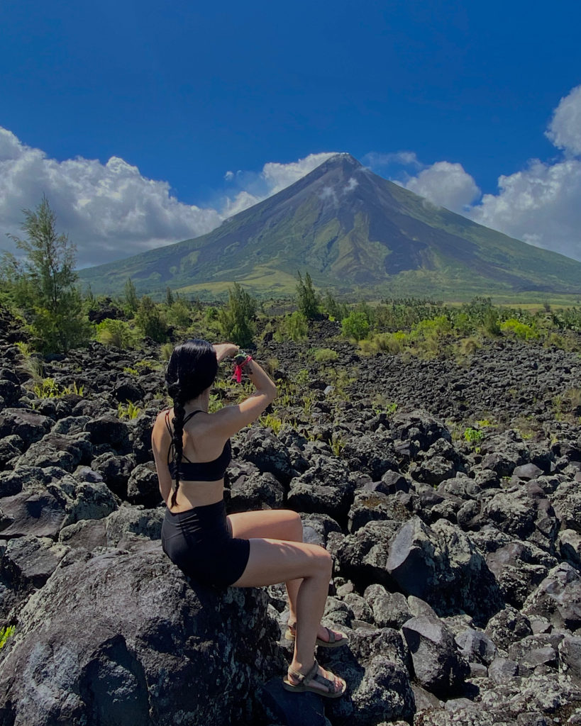 A woman sat on a huge piece of lava with a spectacular view of a cone shaped volcano.