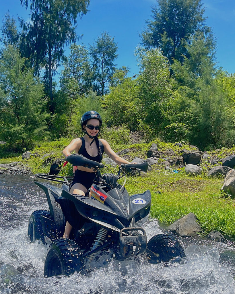 A girl on an ATV driving through a stream surrounded by greenery.