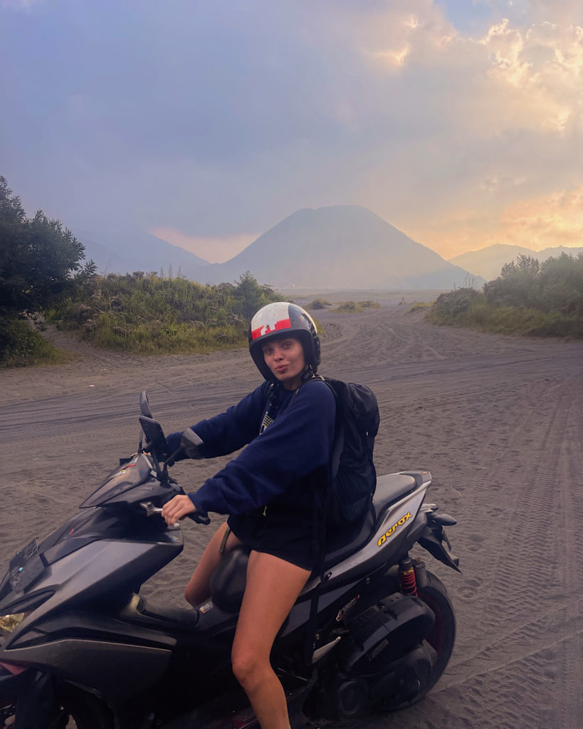 A woman driving a motorbike across black sand in front of Mount Bromo.