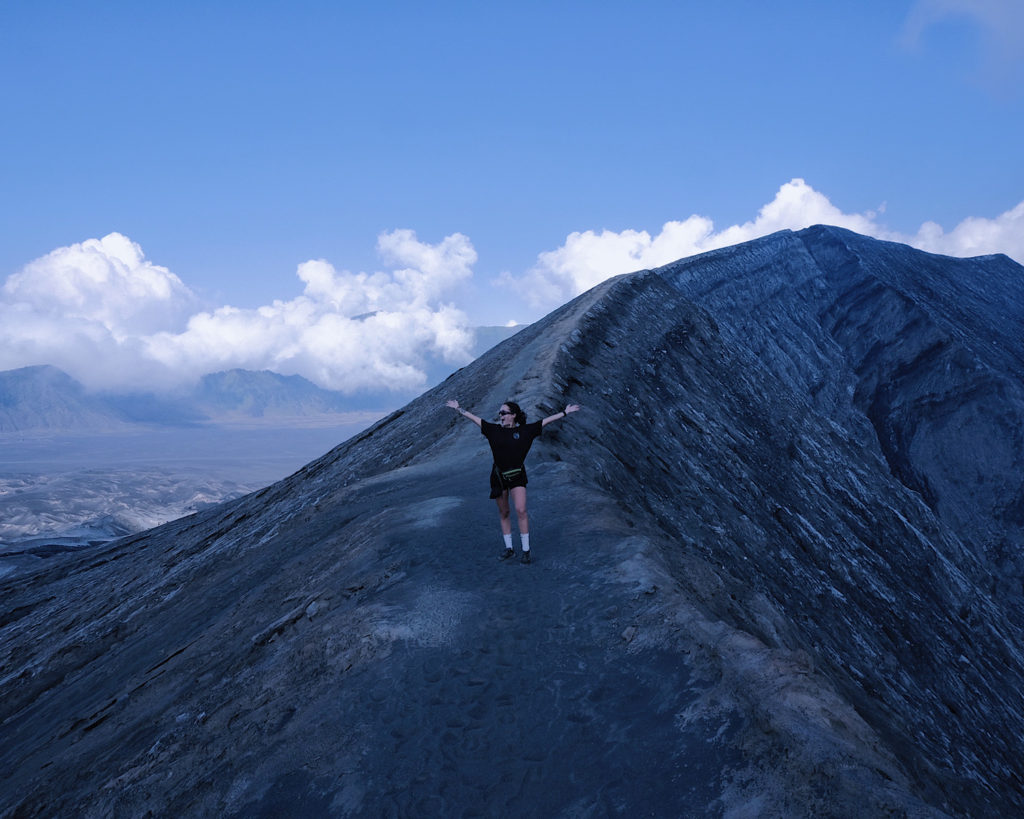 A woman in a black t-shirt standing on the rim of a volcano crater.