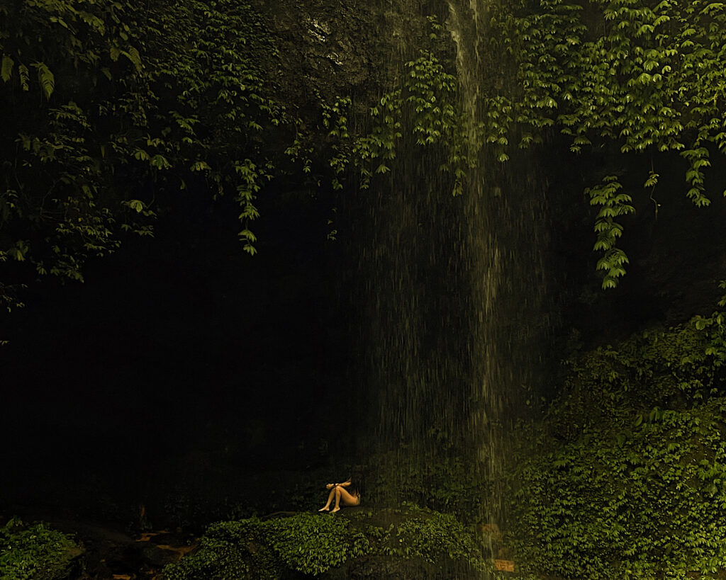 A serene scene of a woman sitting behind a waterfall surrounded by greenery.