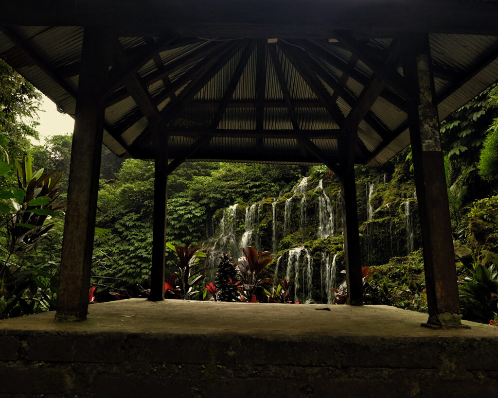 A wooden gazebo situated in front of a magical waterfall surrounded by pretty plantations.