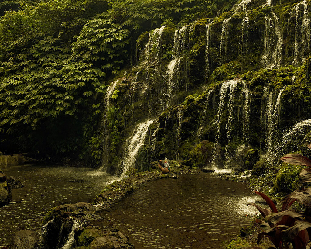 A woman sat in the cool water beneath a tranquil flowing waterfall in the jungle.