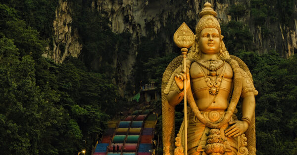 A tall statue of Lord Murugan standing majestically in front of a cave with colourful steps leading to its entrance.