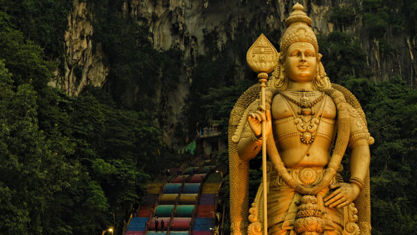 A tall statue of Lord Murugan standing majestically in front of a cave with colourful steps leading to its entrance.