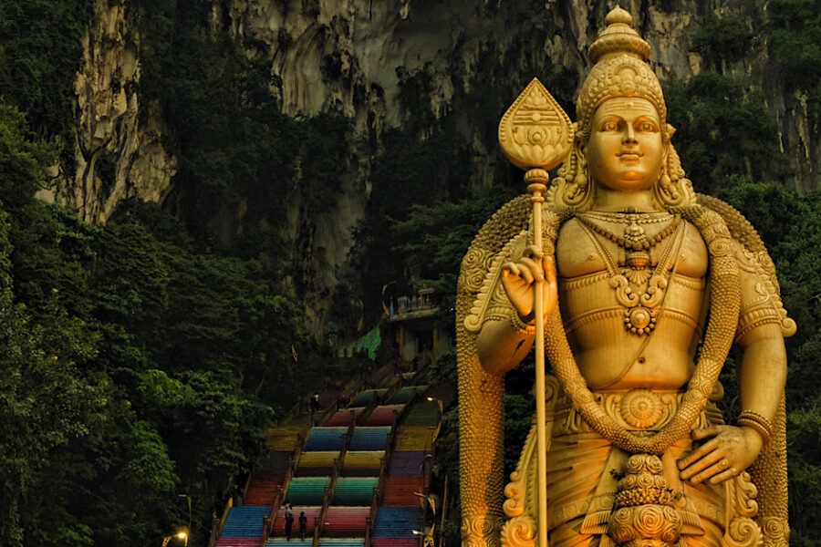 A tall statue of Lord Murugan standing majestically in front of a cave with colourful steps leading to its entrance.