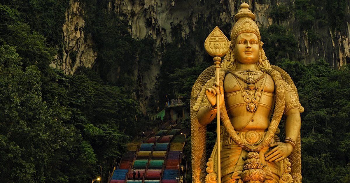 A tall statue of Lord Murugan standing majestically in front of a cave with colourful steps leading to its entrance.