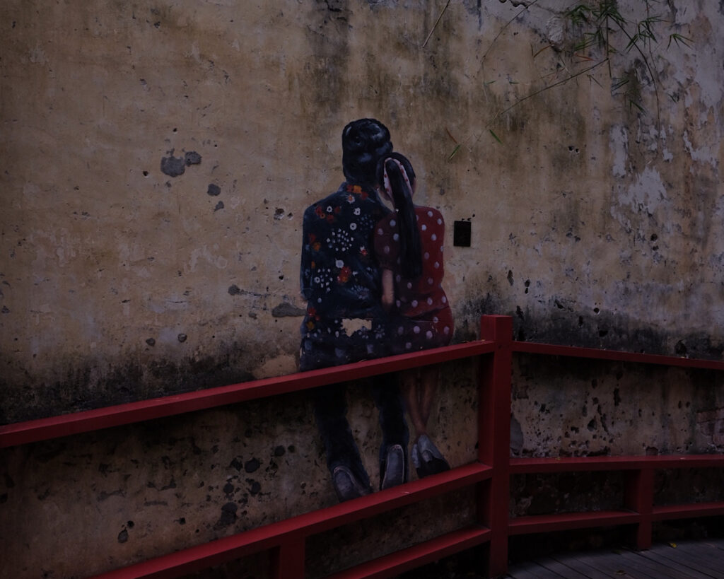 Street art of a man and a woman embracing one another on the wall next to a bridge, giving the illusion that they are seated on the railing.